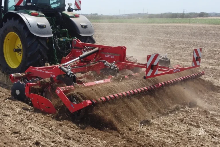 Déchaumeur rouge SIPTEC en fonctionnement dans un champ, attelé à un tracteur agricole moderne.