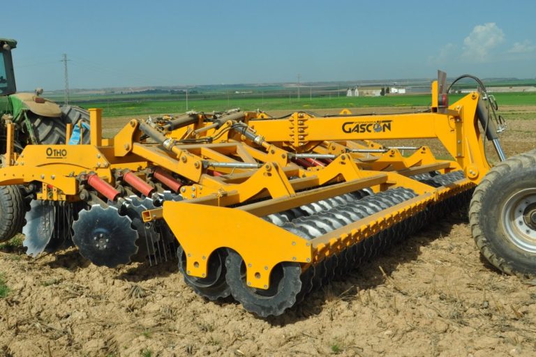 Outil de travail du sol jaune GASCON au repos dans un champ sec, montrant des rouleaux et des dents.