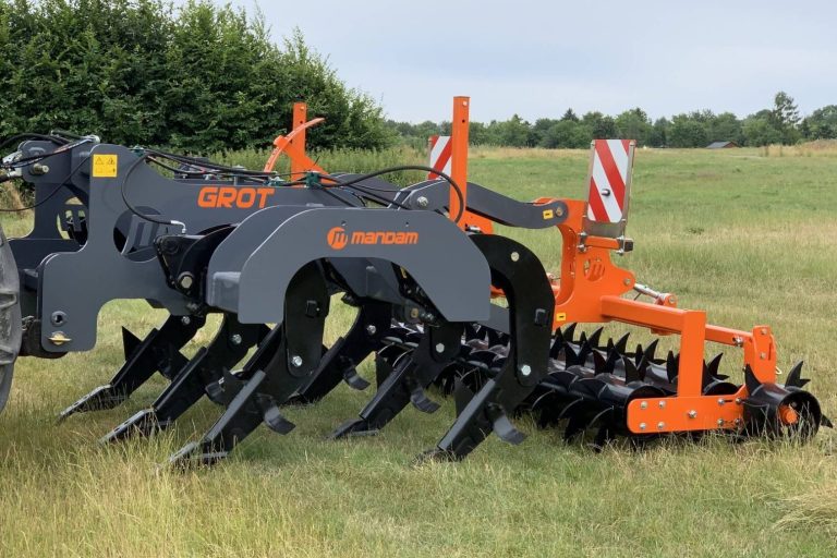 Décompacteur Mandam gris foncé et orange équipé d'un rouleau à pointes, stationné dans l'herbe.