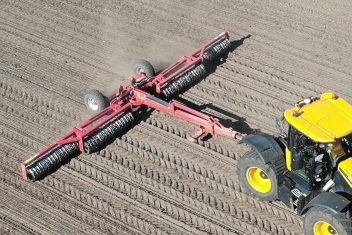 Tracteur attelé à un large rouleau noir et rouge Brix en pleine action pour réamorcer la capillarité après le semis.