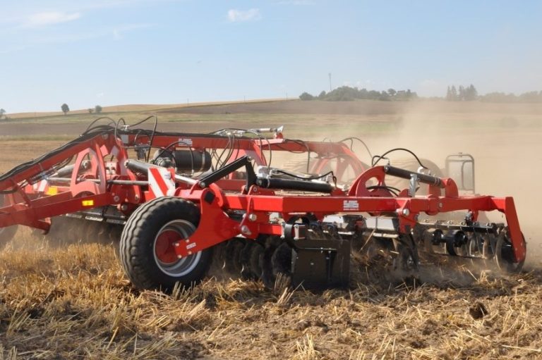Grand déchaumeur rouge SMS AGRO au travail, soulevant de la poussière dans un champ récolté.