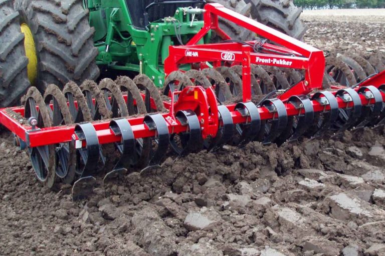 Outil frontal agricole rouge He-Va attelé à l'avant du tracteur pour pré-émietter les mottes avant le passage des roues.
